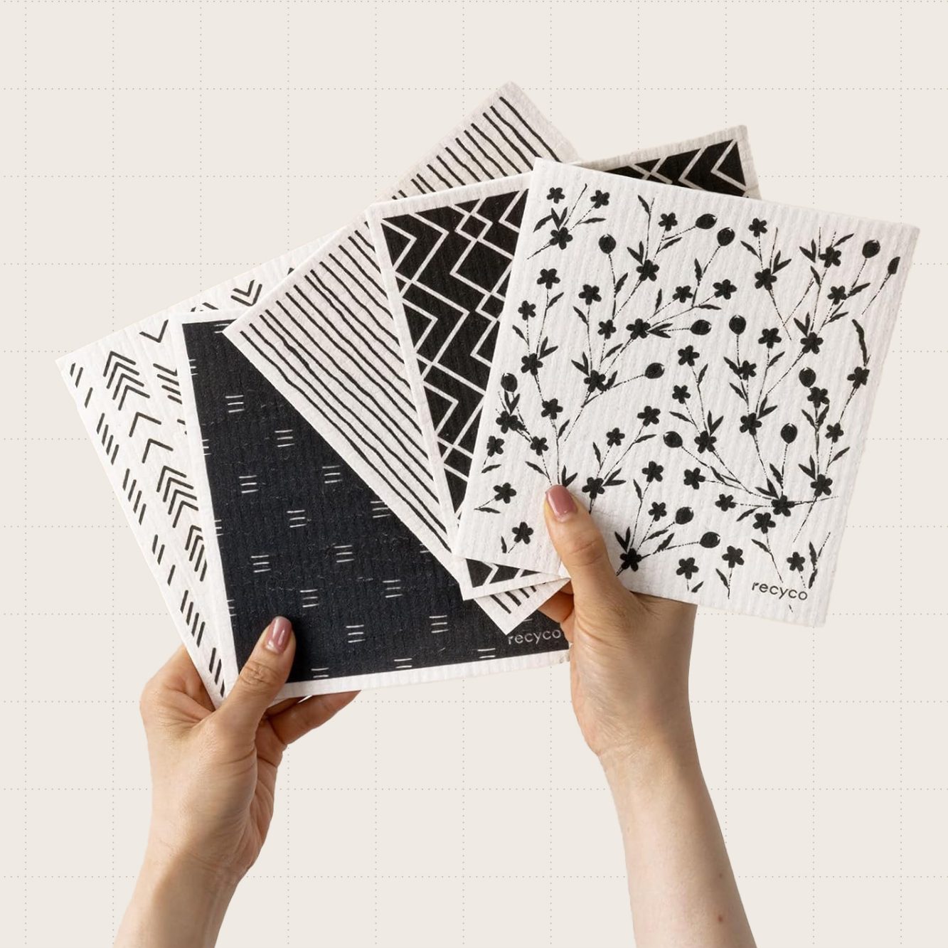 Person's hands holding up a variety pack of black and white Swedish dish cloths with floral and geometric patterns on them.