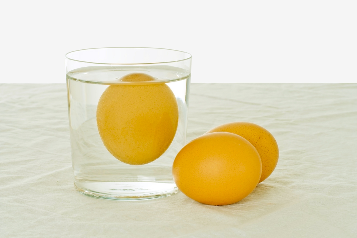 An egg floating in a clear glass of water. Beside the glass are two additional eggs.
