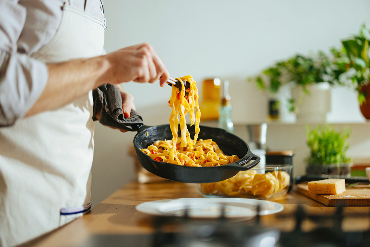Anonymous man cooking pasta in a cast iron skillet and wearing an apron.