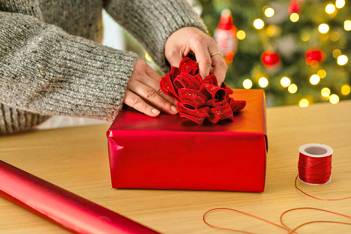 Woman wrapping a gift in red wrapping paper with a glittery bow. In the background is a twinkling Christmas tree.