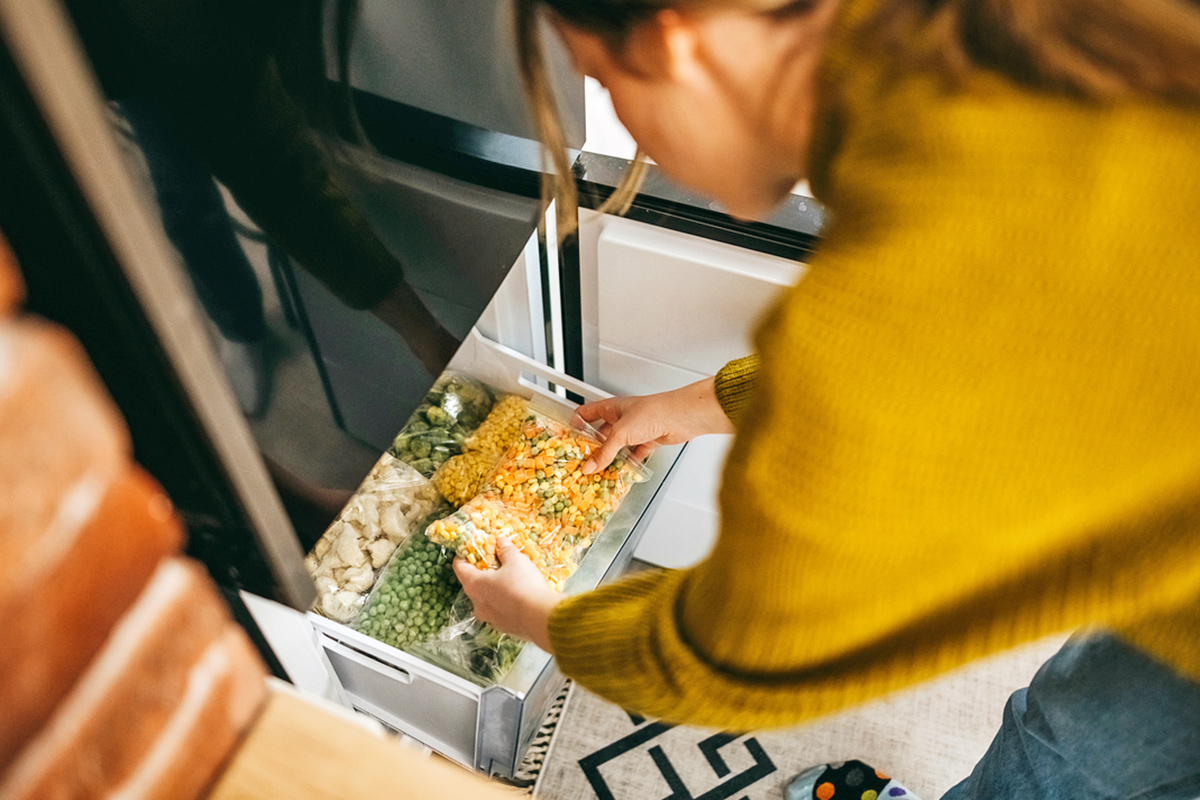 Woman in a sweater placing a bag of frozen corn, carrots, and peas into a freezer drawer.