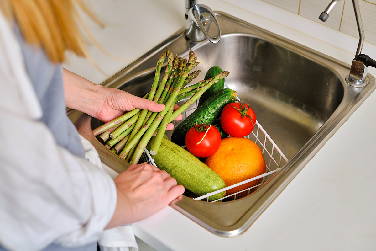 Anonymous woman adding fruits and vegetables to a white wire bin in a stainless steel sink.
