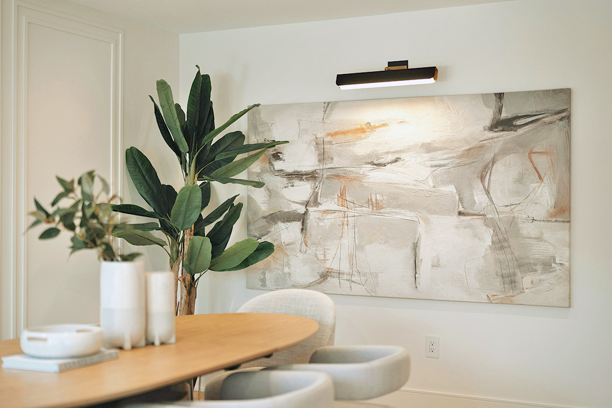 A large abstract painting on a dining room wall that's illuminated by a picture light.