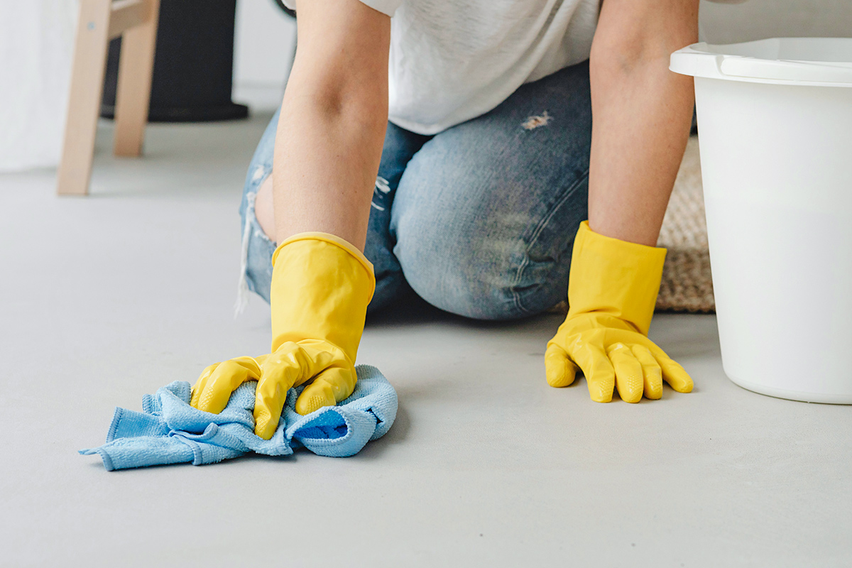 Anonymous woman wearing yellow cleaning gloves scrubbing the floor on her hands and knees. She's using a microfiber cloth and next to her is a white bucket.