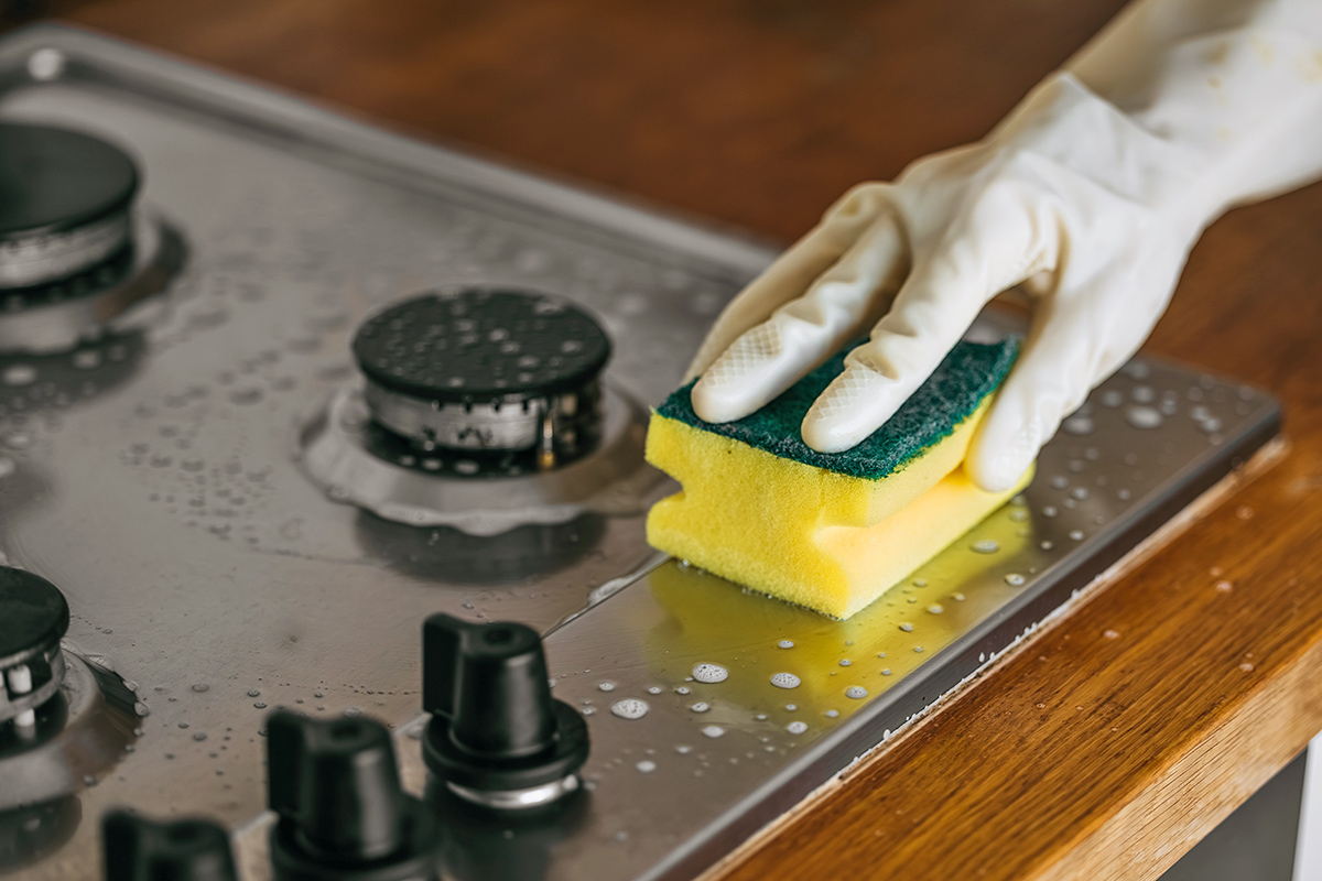 Anonymous person wearing white cleaning gloves scrubbing a stainless steel cooktop with a sponge and cleaning solution.