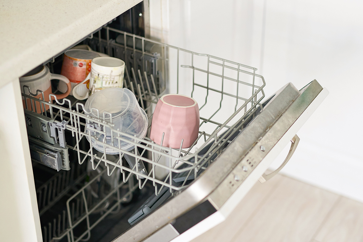 An open dishwasher with the top rack pulled out, revealing several mugs and a food storage container.
