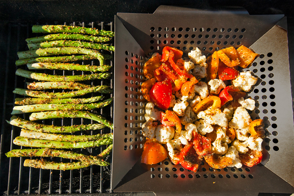 An overhead shot of asparagus on grill grates and diced peppers and cauliflower in a perforated grill basket.